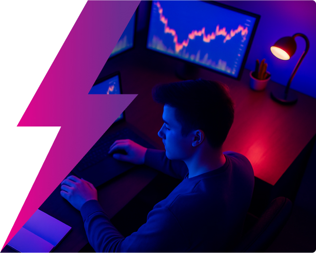 Young professional in their home office with multiple monitors trading. Shot from above with a birds eye view. Dark room with neon lights and the light from the monitor shining on the individual.